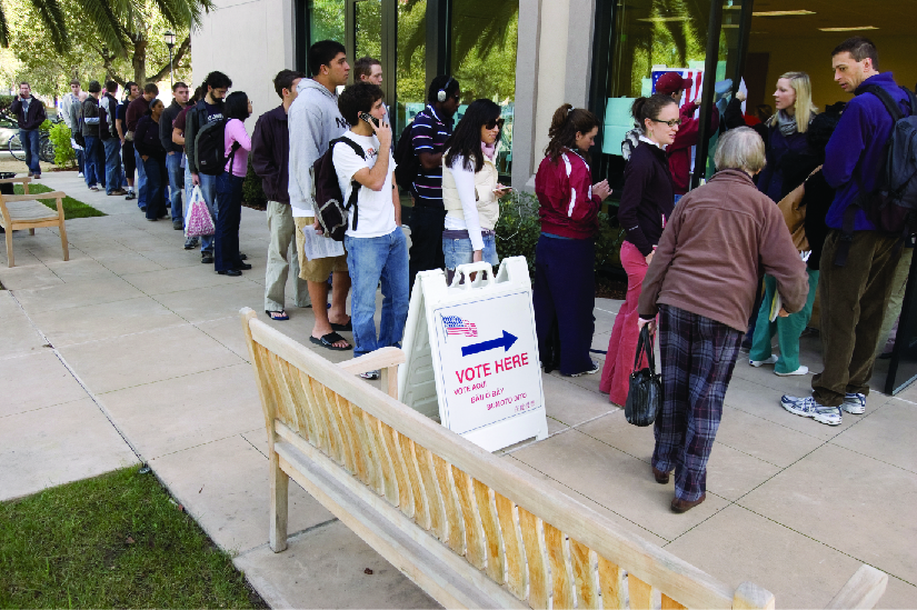 An image of several people standing in line outside of a building. A sign near the front of the line and the building entrance reads “Vote Here”.