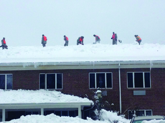An image of several people standing on top of the roof of a brick building. The roof is covered in snow.