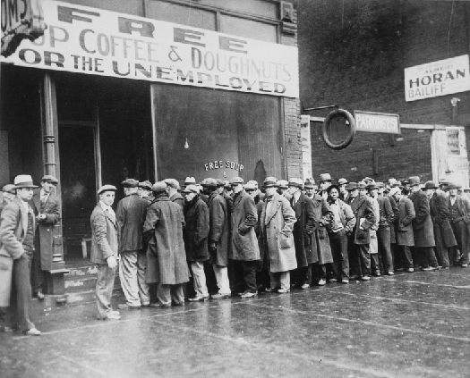 Photo shows a line of people in long coats and hats standing in line outside a building with a sign that states “Free Cup Coffee & Doughnuts for the Unemployed”.